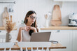 © Iona - Focused young woman with glasses working on laptop at a minimalist kitchen table, her expression one of concentration and determination, surrounded by calm, neutral tones