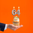 Hand holding birthday cupcake with number 64 candle - background orange