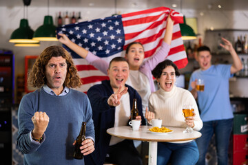 Poster - Emotional company of young adult sports fans supporting American team with state flag while resting in bar with beer