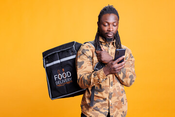 African american courier tracking customer address on smartphone before delivering order during lunch time. Deliveryman holding thermal backpack full with takeaway food over yellow background