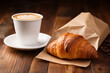 White cup of coffee and croissant in paper bag on wooden background