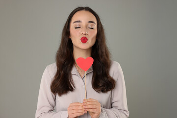 Wall Mural - Beautiful young woman with paper heart sending air kiss on grey background