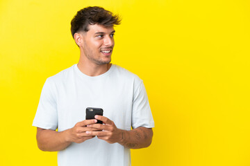 Sticker - Young caucasian handsome man isolated on yellow background using mobile phone and looking up