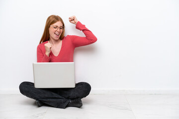 Wall Mural - Young redhead woman with a laptop sitting on the floor isolated on white background celebrating a victory