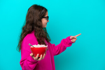 Little girl holding a bowl of cereals isolated on blue background pointing to the side to present a product