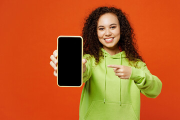 Wall Mural - Young woman of African American ethnicity wears green hoody casual clothes hold use point finger on mobile cell phone with blank screen area isolated on plain red orange background. Lifestyle concept.