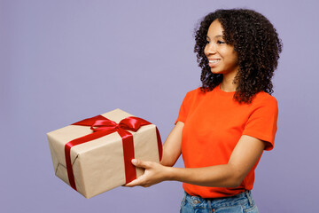 Wall Mural - Side view little kid teen girl of African American ethnicity wear orange t-shirt hold in hand present box with gift ribbon bow isolated on plain pastel purple background. Childhood lifestyle concept.