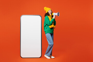 Wall Mural - Full body side view young woman wear green knitted sweater yellow hat scarf big huge blank screen mobile cell phone smartphone with area scream in megaphone isolated on plain orange red background.