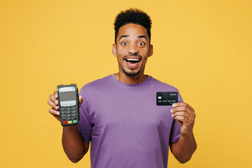 Wall Mural - Young man of African American ethnicity he wears purple t-shirt casual clothes hold wireless bank payment terminal to process acquire credit card isolated on plain yellow background Lifestyle concept