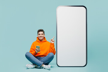 Wall Mural - Full body young man wears orange hoody casual clothes point finger on big huge blank screen mobile cell phone with area use smartphone isolated on plain blue cyan color background. Lifestyle concept.