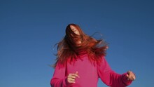 Happy Attractive Young Red Hair Woman In Pink Sweatshirt Having Fun, Dancing, Jumping Outdoors On Blue Sky Backdrop, Bottom View. Girl Moves Her Head With Long Hair To Sides. Freedom And Youth Concept