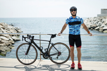 Wall Mural - Fit man standing next to the sea with a cycle wearing helmet and sunglasses. Cyclist leaning against a balcony with a bike wearing sport clothes near to water.