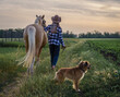 young blonde girl in a hat and a plaid shirt walks with a horse and dog on a farm in the village