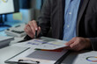 © crizzystudio - Young Asian businessman holds a pen and points to a chart graph on a document to analyze market data, finance, balance sheet, net profit, taxes to plan an online sales strategy startup business idea.