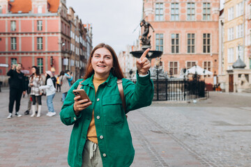 Wall Mural - Happy cheerful young woman with phone walking on city street checks her smartphone. Redhead girl with smart phone pointing finger on city street. Traveling Europe in autumn. Gdansk, Poland