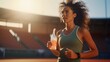 © Ahmad  - photo of a latin female athlete running on the track while holding a drinking bottle in her hand, sweating while exercising, blurred stadium background