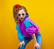 Fashion DJ girl in colorful trendy jacket from 90s. Cool Teenager shows strong face at the disco party of 80s vibes. Fashionable smiling young model on yellow color background.