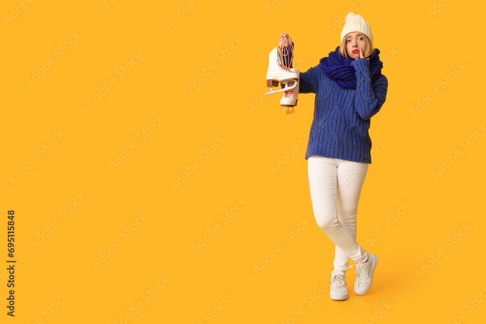 Sad young woman with ice skates on yellow background