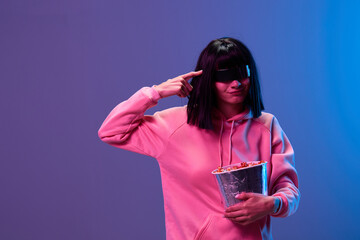 Wall Mural - Funny confused brunet woman in pink hoodie trendy specular sunglasses with popcorn hold finger on temple posing isolated in blue violet color light background. Neon party Cinema concept. Copy space