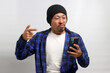 An enraged Asian man, dressed in a beanie hat and casual shirt, points at his mobile phone while visibly reacting to bad news, showing intense anger and fury, while standing against white background