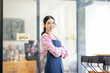 © David - Portrait of happy waitress standing at restaurant entrance. SME entrepreneur young business asian woman attend new customers near door. Smiling small business owner showing open sign in her shop.
