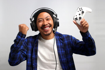 An enthusiastic Asian gamer, adorned in a beanie hat, casual shirt, and headphones, celebrates victory while playing a video game on a console with a gamepad or joystick, isolated on white background.