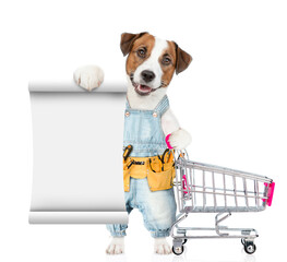 Poster - Funny Jack russell terrier puppy wearing denim overalls with tool belt shows empty list and holds the shopping cart. isolated on white background