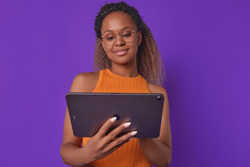 Young cute optimistic African American woman with electronic tablet in hands uses gadget to surf internet on social networks and view photos of boyfriend or parents stands posing on purple background.