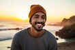 © Markus Schröder - Portrait of a smiling indian man in his 20s sporting a trendy beanie against a beautiful beach sunset. AI Generation