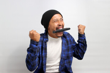 Funny young Asian man, dressed in a beanie hat and casual shirt, bites the TV remote in frustration while feeling angry and frustrated while watching TV, standing against a white background