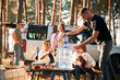 © standret - Sitting by the table with food, against car. Group of friends are together in the forest