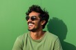 © Markus Schröder - Portrait of a smiling indian man in his 20s wearing a trendy sunglasses against a soft green background. AI Generation