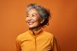 © Markus Schröder - Portrait of a blissful asian woman in her 80s sporting a breathable hiking shirt against a soft orange background. AI Generation