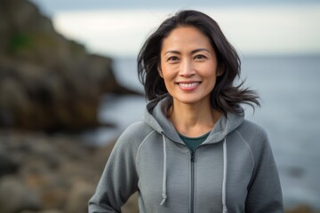 Wall Mural - Portrait of a smiling asian woman in her 40s wearing a zip-up fleece hoodie against a rocky shoreline background. AI Generation