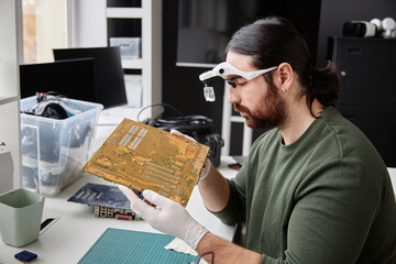 Side view portrait of bearded repairman inspecting circuit board with magnifying glasses in computer repair shop, copy space