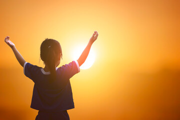 Wall Mural - Silhouette of little girl raising hands on sunset sky background.