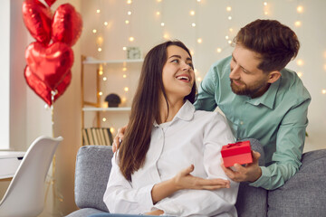 Wall Mural - Portrait of man holding red present box in his hand hugging and congratulating pretty woman on holiday sitting on sofa at home. Happy couple in love. Care, congratulations and Valentines day concept.