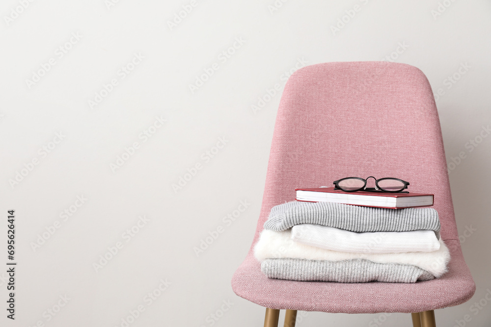 Stack of folded sweaters with book and eyeglasses on pink chair against white background