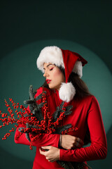 Wall Mural - Beautiful young woman in Santa hat holding Christmas branches with rowan on green background