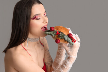 Wall Mural - Beautiful young woman holding burger with Christmas balls and fir branches on grey background