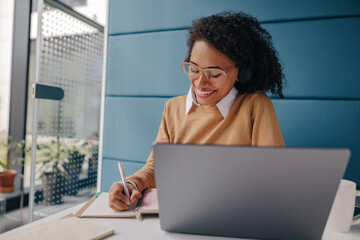 Wall Mural - Smiling female analyst in eyeglasses working on laptop and making notes while sitting in cozy office