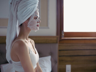 Young woman lying on bed with moisturizing beauty face mask and white towel on her head after shower, lifestyle home skincare, smile.