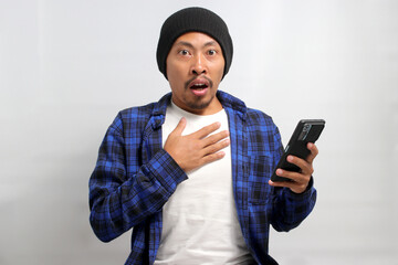 An astonished Asian man, dressed in a beanie hat and casual shirt, reacts with surprise to an ecommerce special offer discount on his phone while standing against a white background