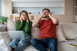 © DimaBerlin - Annoyed unhappy couple sitting on sofa with vexed face expression, looking upward, closing ears because of loud sound, noise from neighboring flat. Emotional stress, discomfort caused by neighborhood