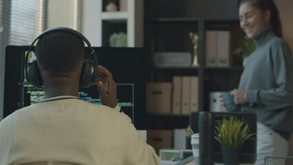 Medium shot of young Caucasian female office worker with cup of coffee in hands talking with Black male program developer sitting in front of desktop computer with program code on screen