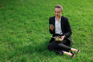 Wall Mural - Lunch time. Happy businesswoman with container of salad using smartphone on green grass outdoors, space for text