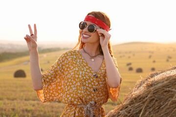 Poster - Happy hippie woman showing peace sign near hay bale in field