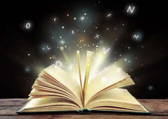 Wall Mural - Open book with magic light and glowing letters flying out of it on wooden table against black background