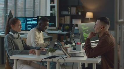 Medium shot of ethnically diverse team of programmers sitting at one table in modern office, talking and coding on their computers