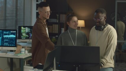 Medium shot of multiethnic programmers team working on new software together standing by desktop computer in modern office and having discussion